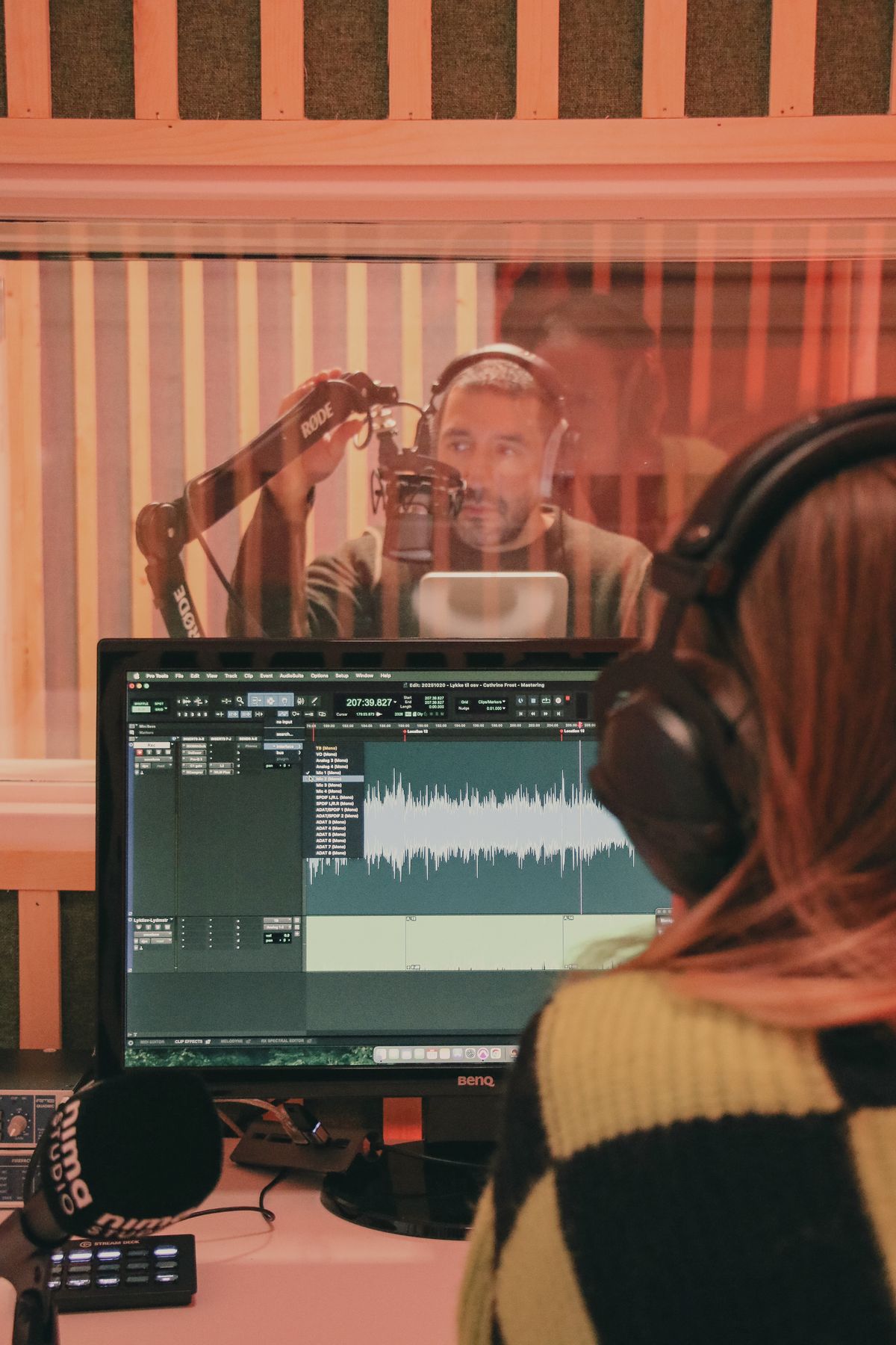 Produsent ved miksepulten mens innleser er i opptak bak glasset